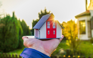 Symbolbild Wohnvorlieben: Hand hält kleines Miniaturhaus vor echtes Einfamilienhaus