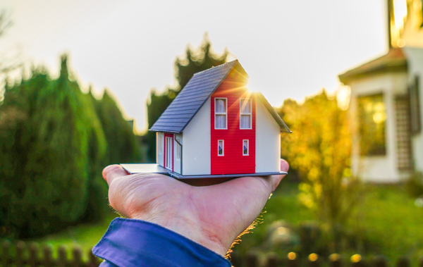 Symbolbild Wohnvorlieben: Hand hält kleines Miniatur-Haus vor echtes Einfamilienhaus