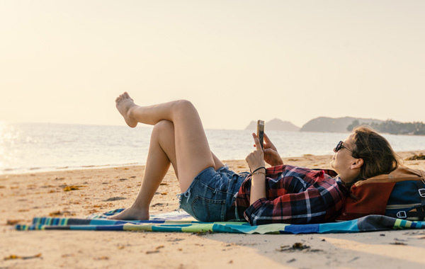 Symbolbild zum Thema Urlaubszeit ist Einbruchzeit: Frau liegt am Strand und bedient Smartphone