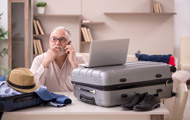 Symbolbild Ferienzeit: Älterer Mann mit gepackten Koffern sitzt am Laptop und telefoniert