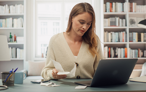 Symbol-Bild Arbeitszimmer: Eine junge Frau sitzt zu Hause am Schreibtisch und schaut auf einen Laptop. Der Schreibtisch und die Regale vermitteln eine Arbeitsplatz-Atmosphäre.