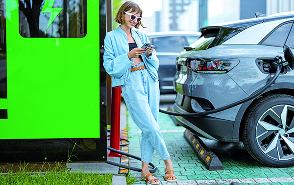 Symbol-Bild Wallbox: Junge Frau lehnt sich an und schaut auf ihr Mobiltelefon, während ihr Elektroauto geladen wird.