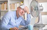 Symbol-Bild: Mann leidet trotz Ventilator und feuchtem Tuch auf der Stirn